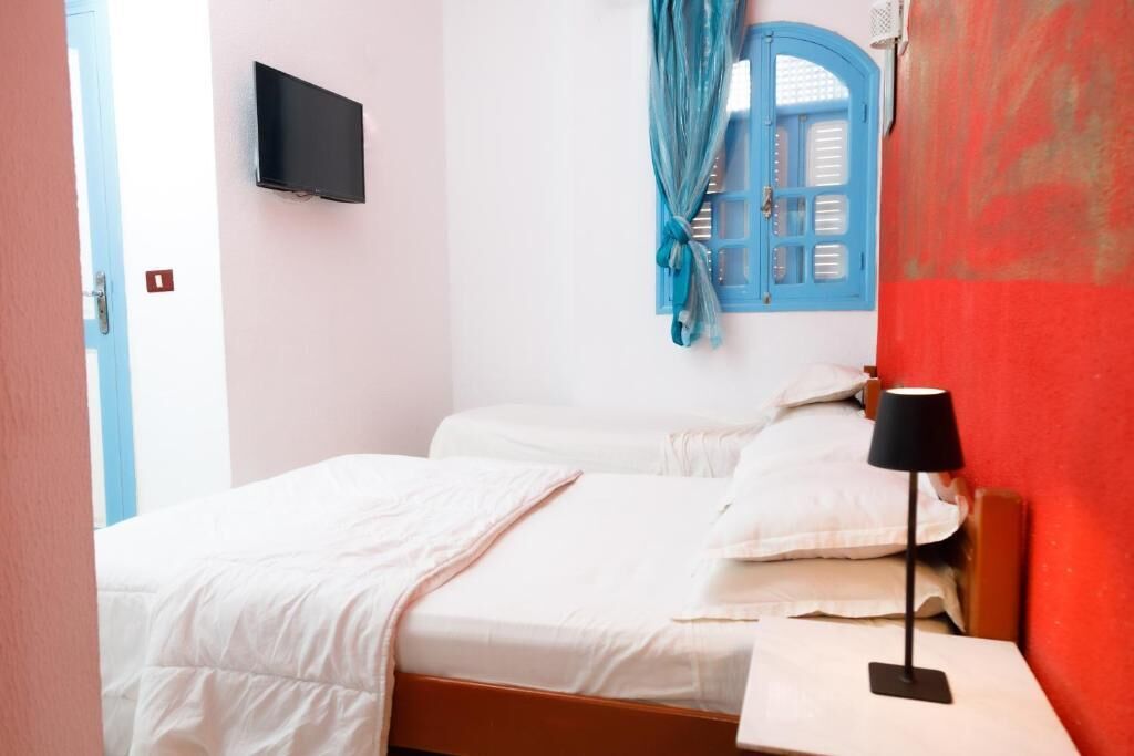 A bedroom with a bed, a bedside table with a lamp, a TV mounted on the wall, and a window with blue shutters.