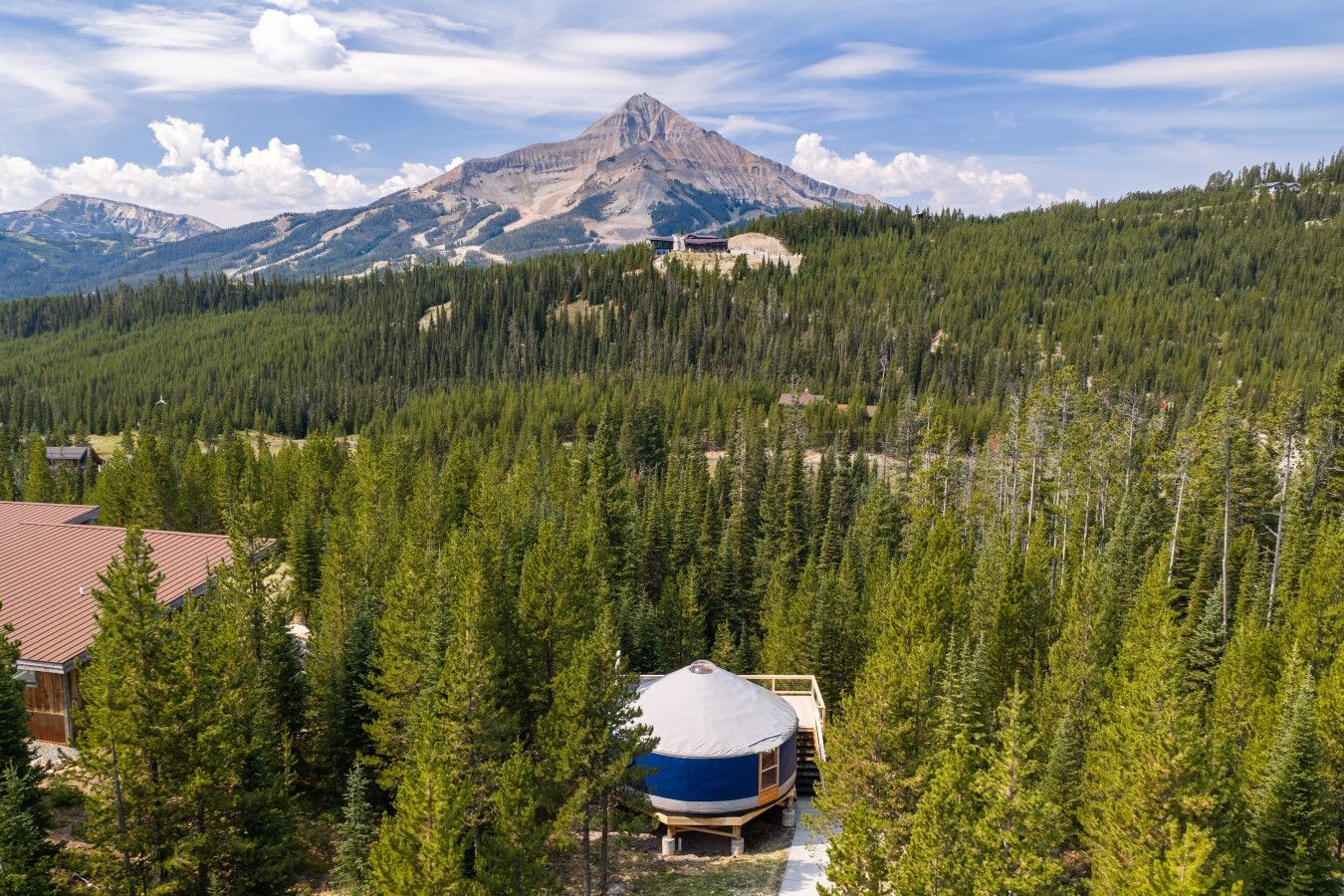 Une yourte avec un toit bleu, entourée d’une forêt dense, et une montagne en arrière-plan.