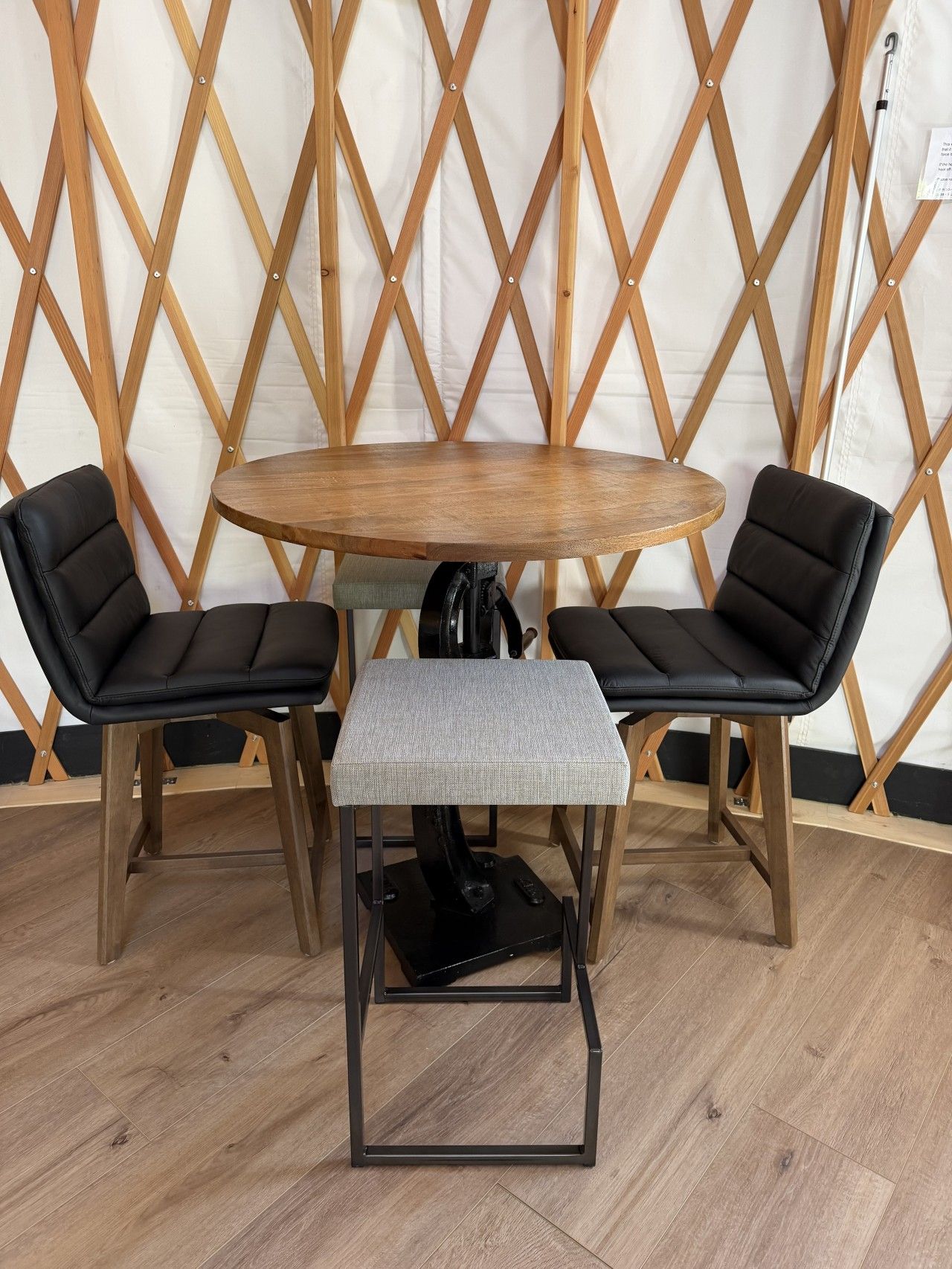 Ein runder Holztisch mit zwei schwarzen Ledersesseln, einem grauen gepolsterten Hocker und einer Holzwand mit Gittermuster im Hintergrund.