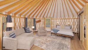 Interior de uma yurtura com sofá, poltrona e cama, vigas de madeira e uma janela.