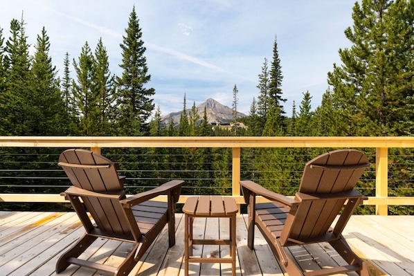 A wooden deck with two Adirondack chairs and a small table, overlooking a forest and a mountain.