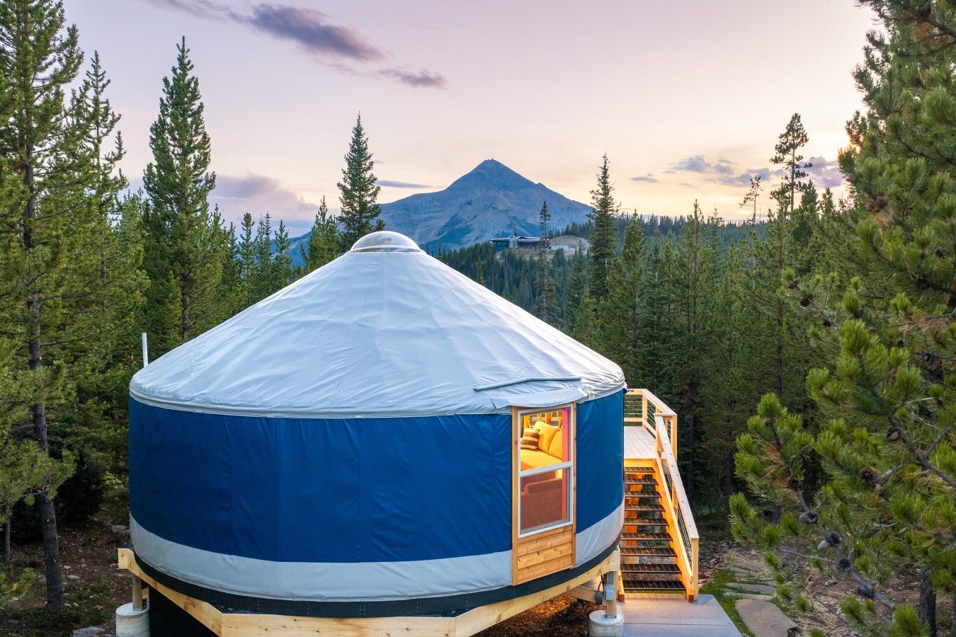 A yurt with a wooden staircase leading to a platform, surrounded by trees and a mountain in the background.