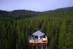 Estructura tipo yurt con techo cónico, rodeada de un bosque denso, elevada sobre pilares y con una terraza de madera con asientos.