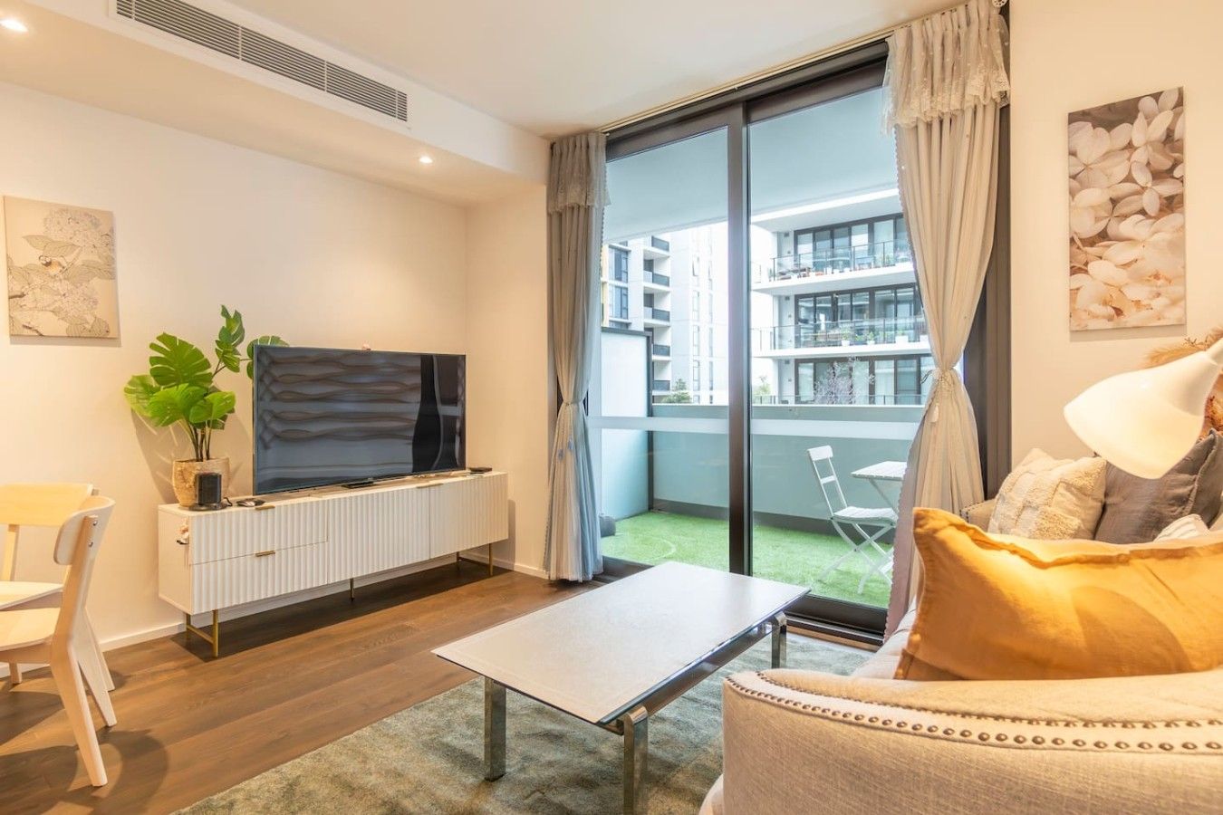A modern living room with a flat-screen TV, a dining table, a sofa, and a balcony with a view of the city.