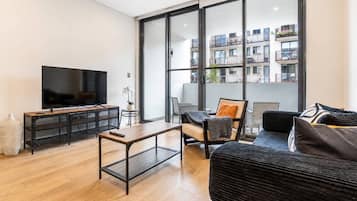 A modern living room with a flat-screen TV, a wooden coffee table, a sofa, and a large window offering a view of a building with balconies.