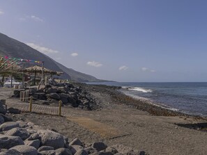 Ein Strand mit einem strohgedeckten Gebäude, einer Holzpromenade und einer felsigen Küstenlinie.