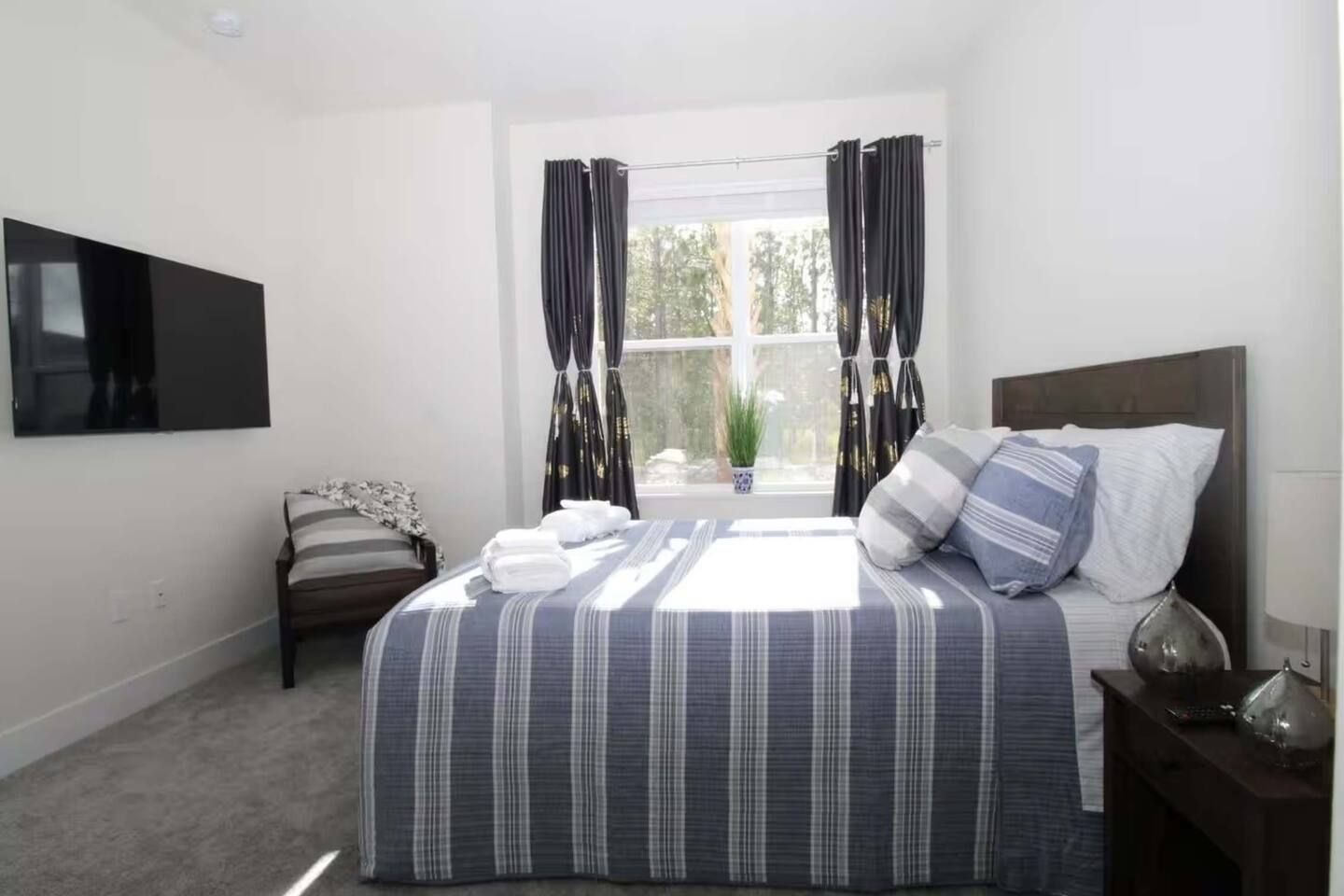 A bedroom with a bed, a TV mounted on the wall, a window with curtains, and a bedside table with a lamp.