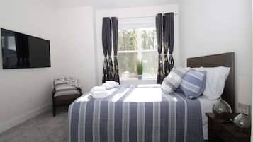 A bedroom with a bed, a TV mounted on the wall, a window with curtains, and a bedside table with a lamp.