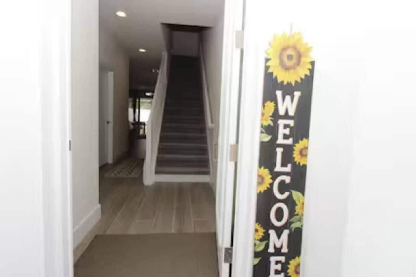 A hallway with a staircase leading to another level, a welcome sign with sunflowers, and a doormat at the entrance.