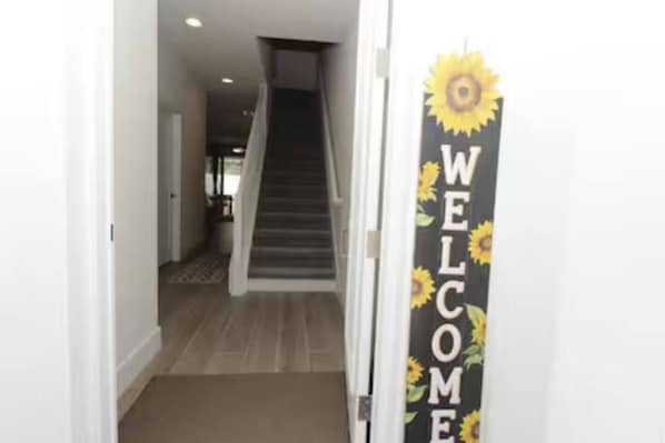 Un pasillo con una escalera que sube a otro nivel, un letrero de bienvenida con girasoles y un felpudo en la entrada.