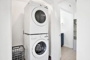 Une machine à laver et un sèche-linge superposés sont installés dans un couloir, avec une porte et un petit espace de rangement en arrière-plan.