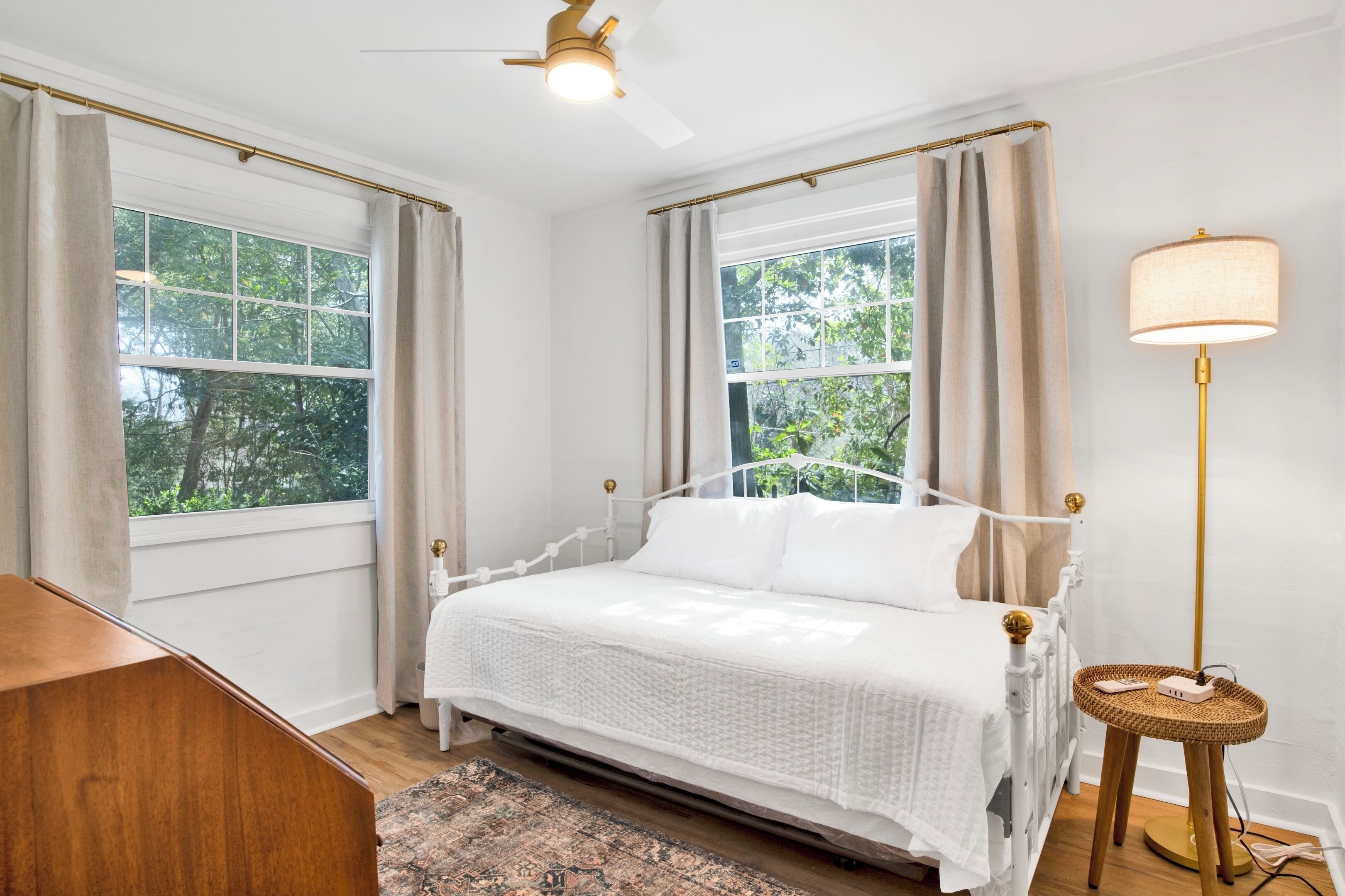 Quarto com cama branca, luminária de chão e vista para árvores pelas janelas.