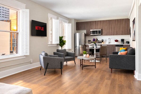 A modern living room with a flat-screen TV, grey sofas, a dining table, and a kitchen in the background. - The Penn Hotel - 2BR Above Nightlife & Top Clubs!! (San Diego)