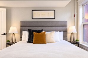 A neatly made bed with a grey upholstered headboard, flanked by two bedside tables with lamps and small potted plants. - The Penn Hotel - 2BR Above Nightlife & Top Clubs!! (San Diego)