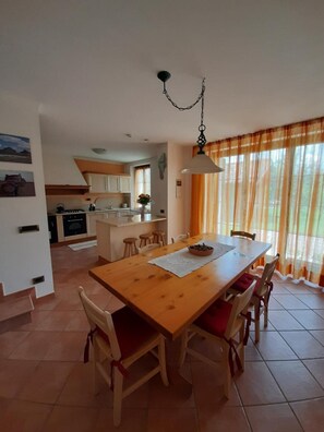 A dining area with a wooden table and chairs, a kitchen in the background, and a window with curtains.