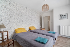 A bedroom with two beds, a wooden nightstand, a lamp, a framed picture on the wall, and a radiator. - Spacious Apartment in St Malo-Paramé with Terrace and Garage (Saint-Malo)