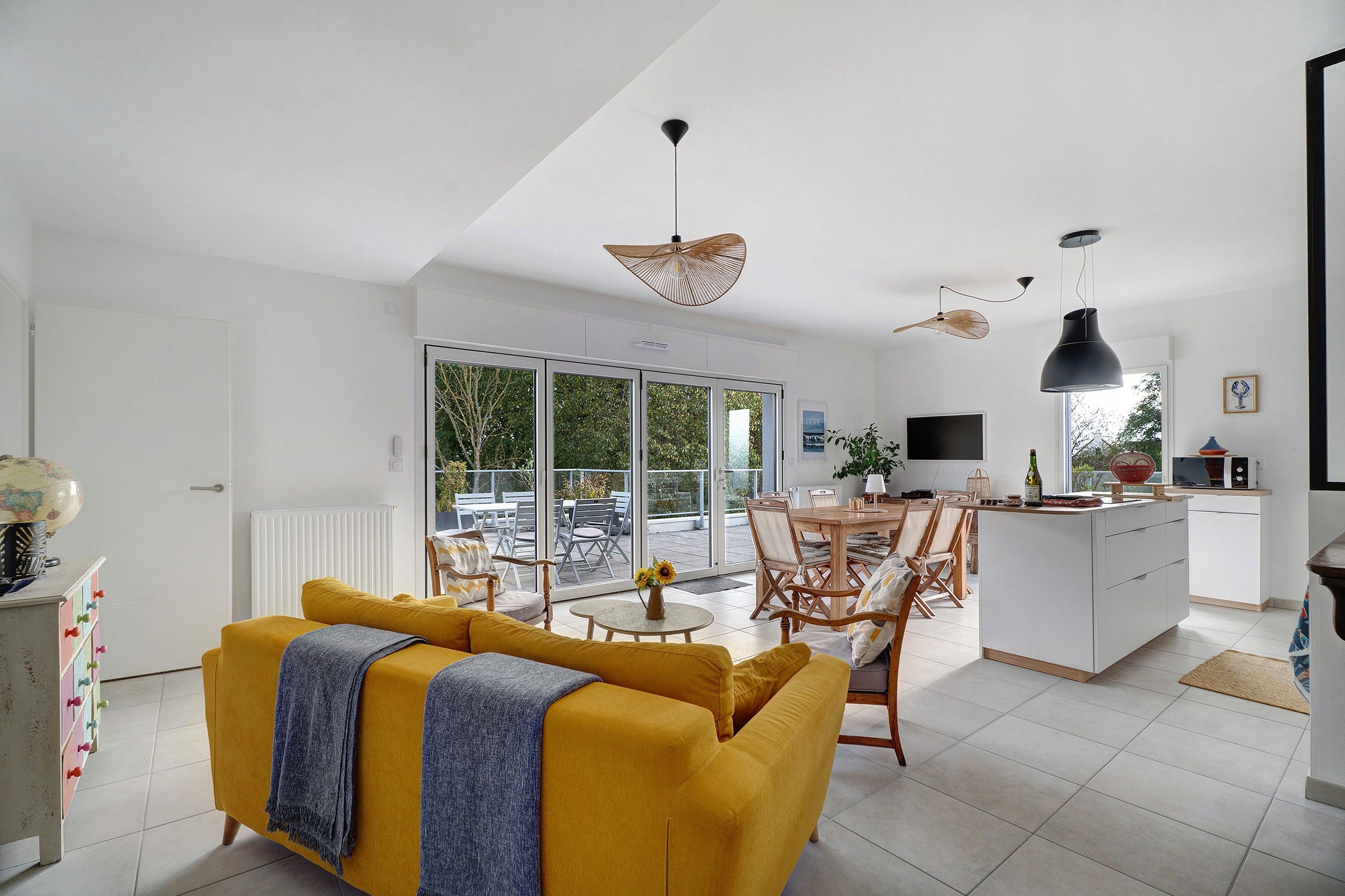 Een moderne woonruimte met een gele bank, eethoek, keuken en grote schuifdeuren naar een balkon.