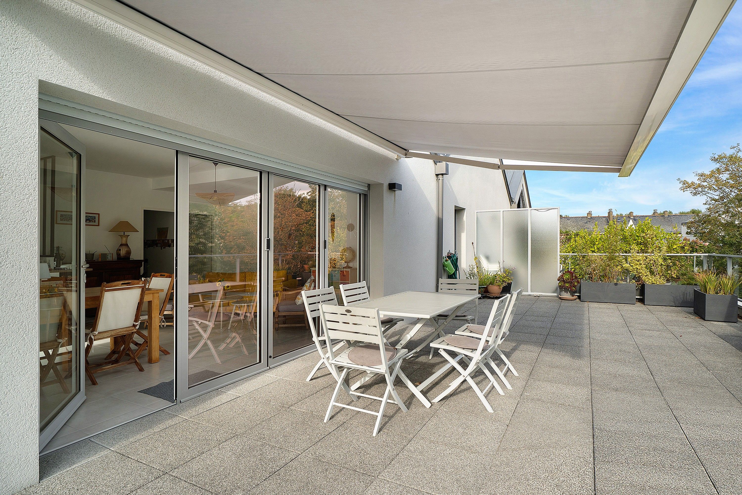 En udendørs terrasse med et bord og stole, en glasdør ind til et rum indendørs, og plantekrukker.