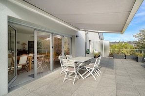 En udendørs terrasse med et bord og stole, en glasdør ind til et rum indendørs, og plantekrukker.