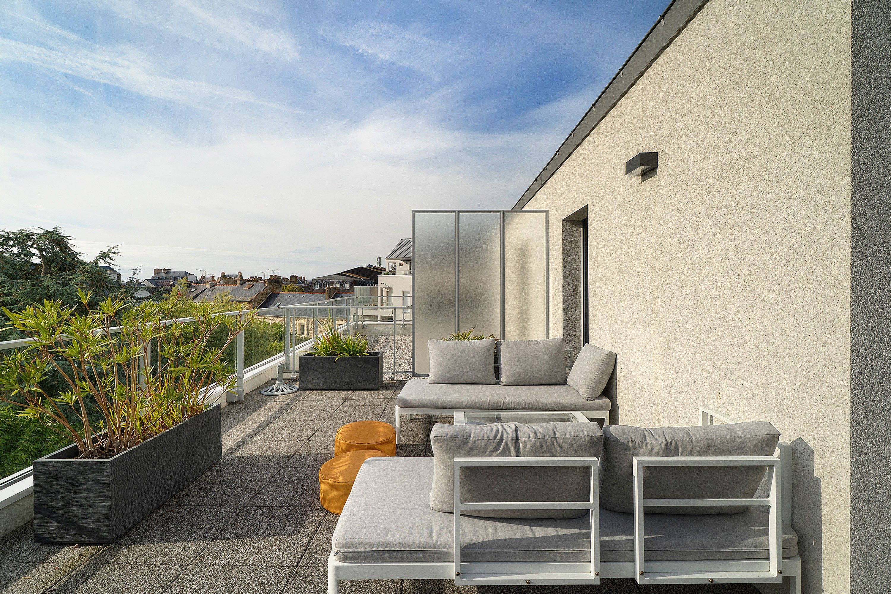 Une terrasse sur le toit dotée d’un espace salon extérieur moderne, comprenant un canapé et un pouf, ainsi qu’une plante en pot.