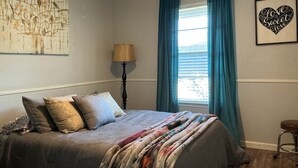Een slaapkamer met een bed, een lamp, een raam met gordijnen en een kunstwerk aan de muur.