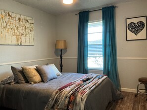 Un dormitorio con una cama, una lámpara, una ventana con cortinas y una obra de arte en la pared.