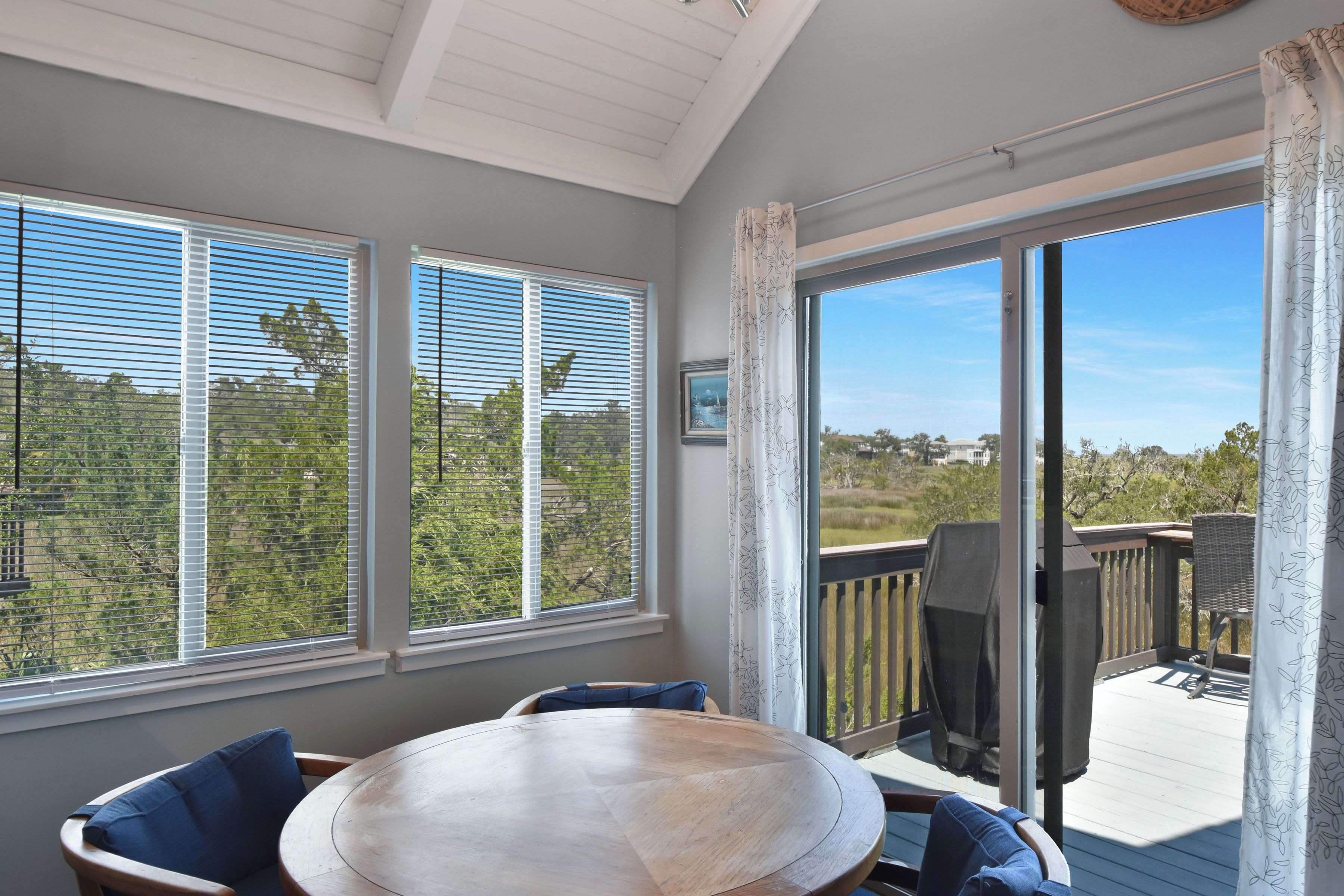A sunroom with a large table and chairs, a sliding glass door leading to an outdoor deck, and a view of greenery and trees.