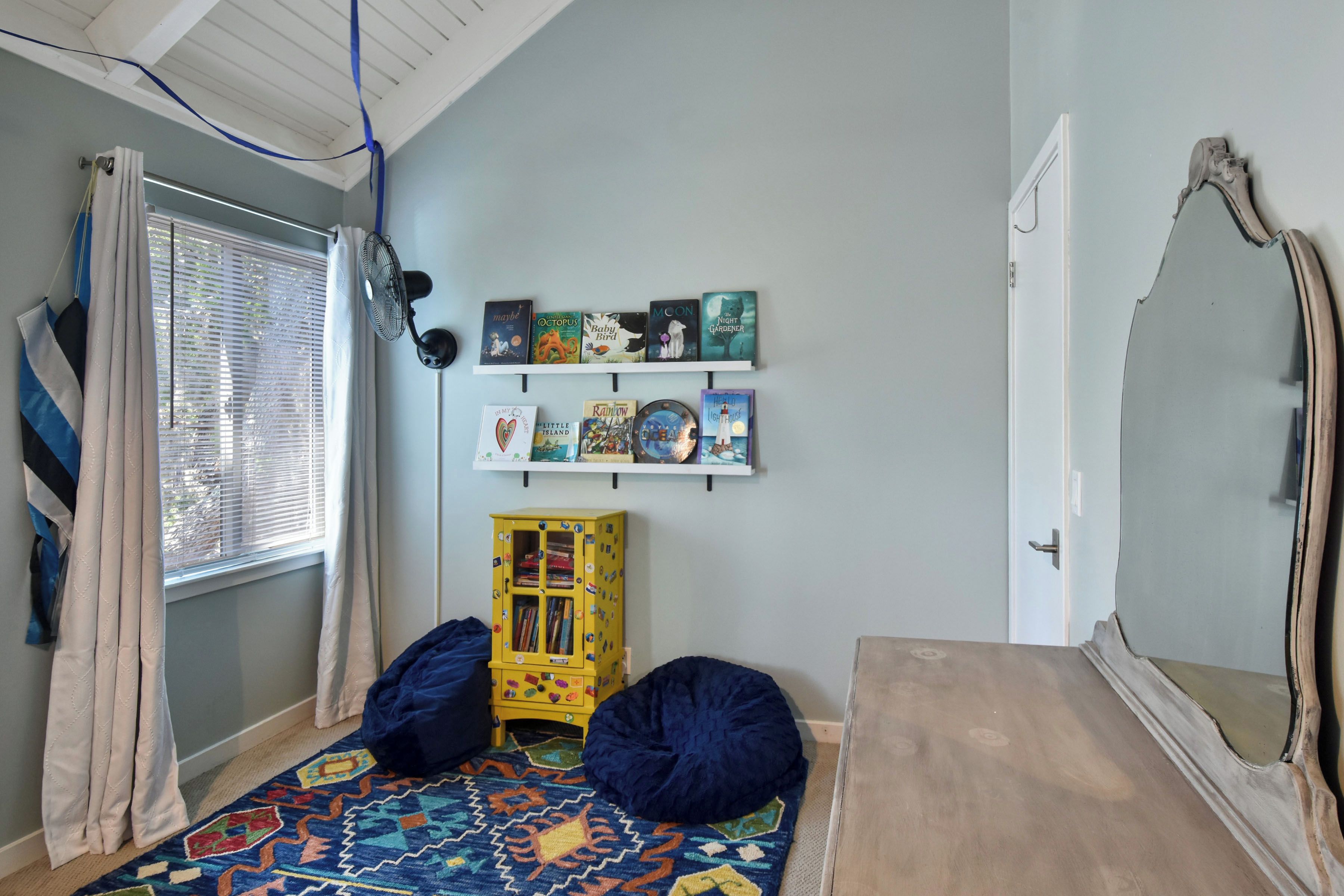 A room with a bed, a window with blinds, a wall-mounted fan, a bookshelf with books, a yellow cabinet, and a mirror.