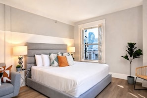 A modern bedroom with a large bed, bedside lamps, a view of the city through a window, and a potted plant. - The Penn Hotel - Walk to Attractions & Sleeps 5!! (San Diego)