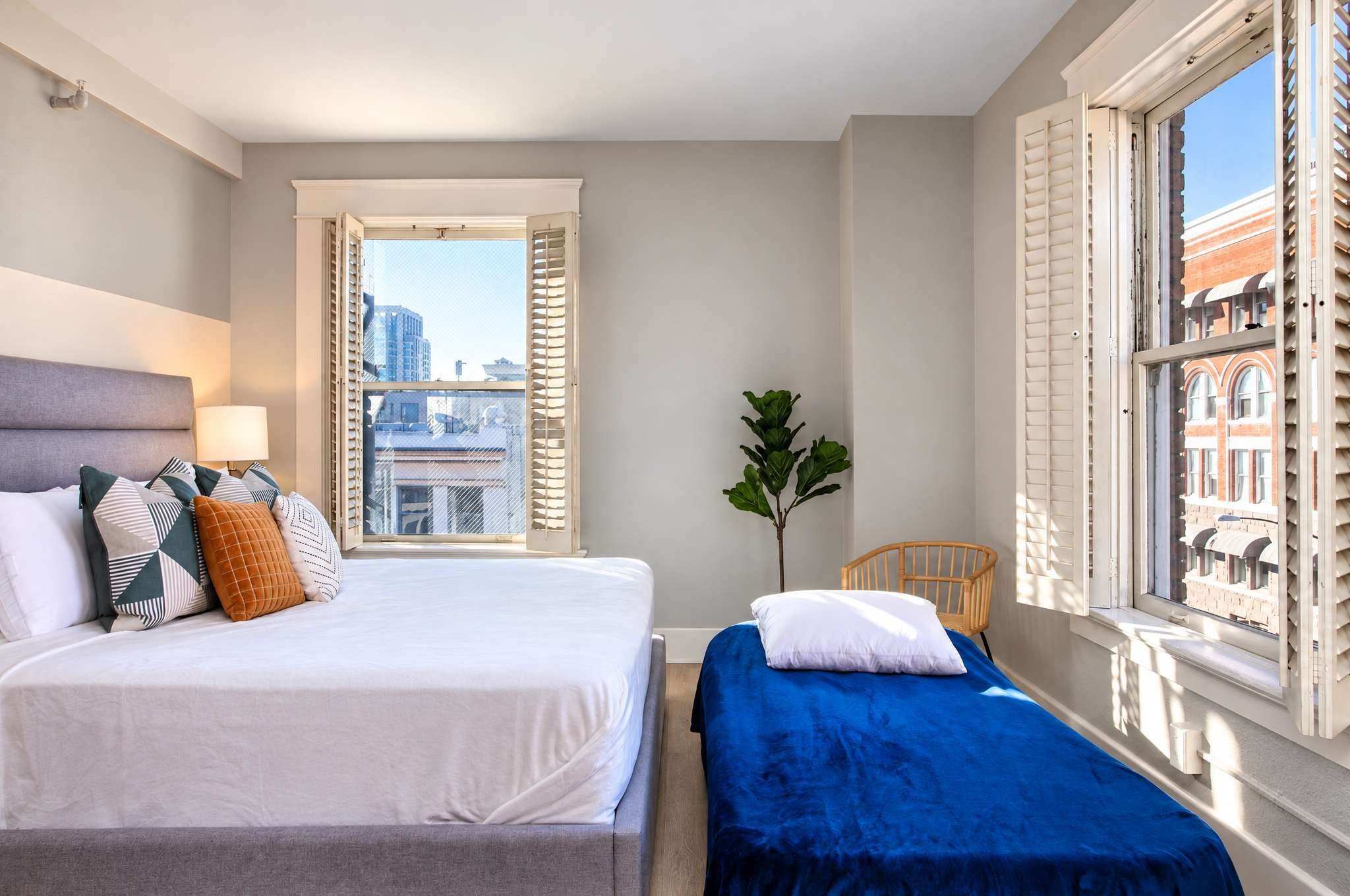 A hotel room with two beds, a window with shutters, a plant, and a view of buildings.