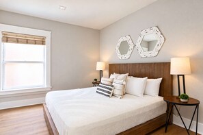 A bedroom with a large bed, two bedside tables with lamps, a window with blinds, and two wall-mounted mirrors. - The Penn Hotel - King & Queen Bed | Gaslamp Life (San Diego)