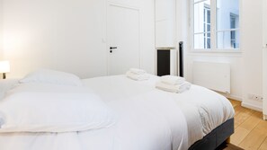 Una cama grande con sábanas blancas, una mesita de noche con una lámpara y una ventana con vistas al exterior.