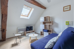 A modern, well-lit room with a sloped ceiling, a television, a small table, a sofa, and a built-in shelving unit. - Le 3 Saint-Blaise - Joli studio à Autun (Autun)