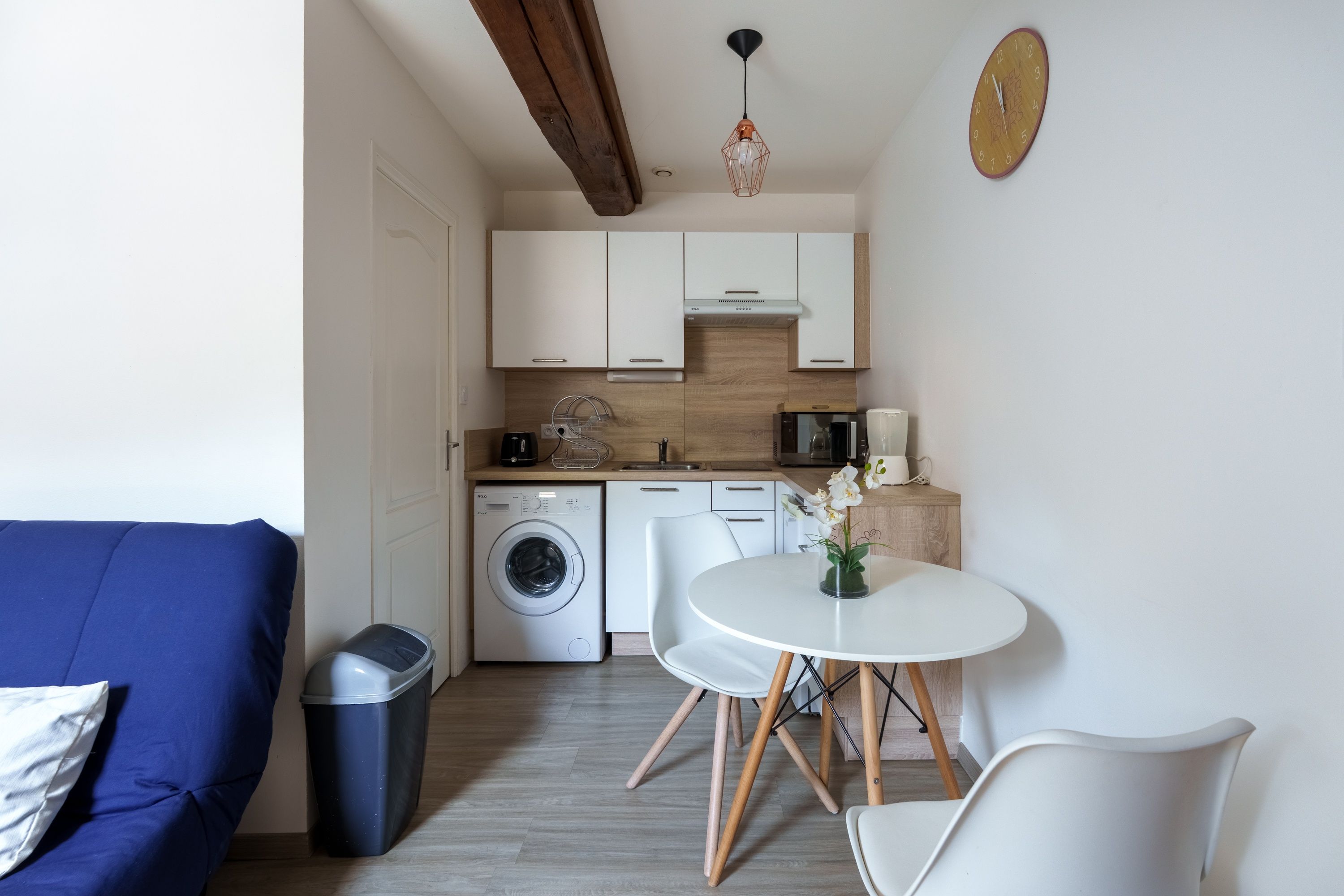Een kleine keuken met een wasmachine, een koffiezetapparaat en een eethoek met een tafel en stoelen.