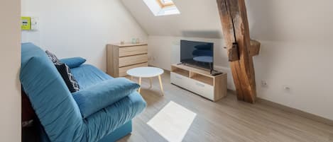Et loftstudie med skrå loft, indbygget tv-møbel, blå sofa, lille hvidt bord og et ovenlys.