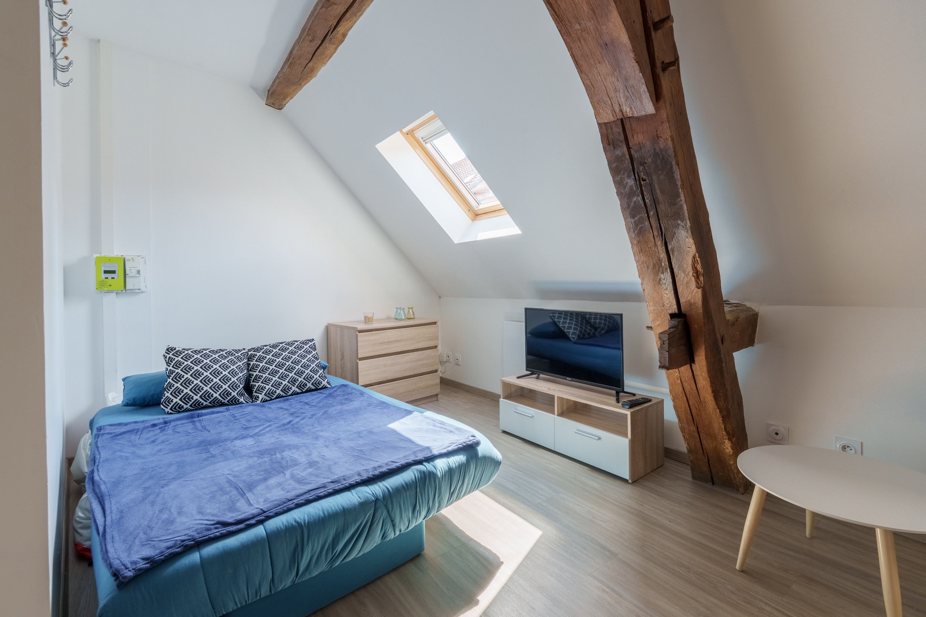 A bedroom with a sloped ceiling, a bed with a blue bedspread, a wooden dresser, a TV on a wooden stand, and a skylight.