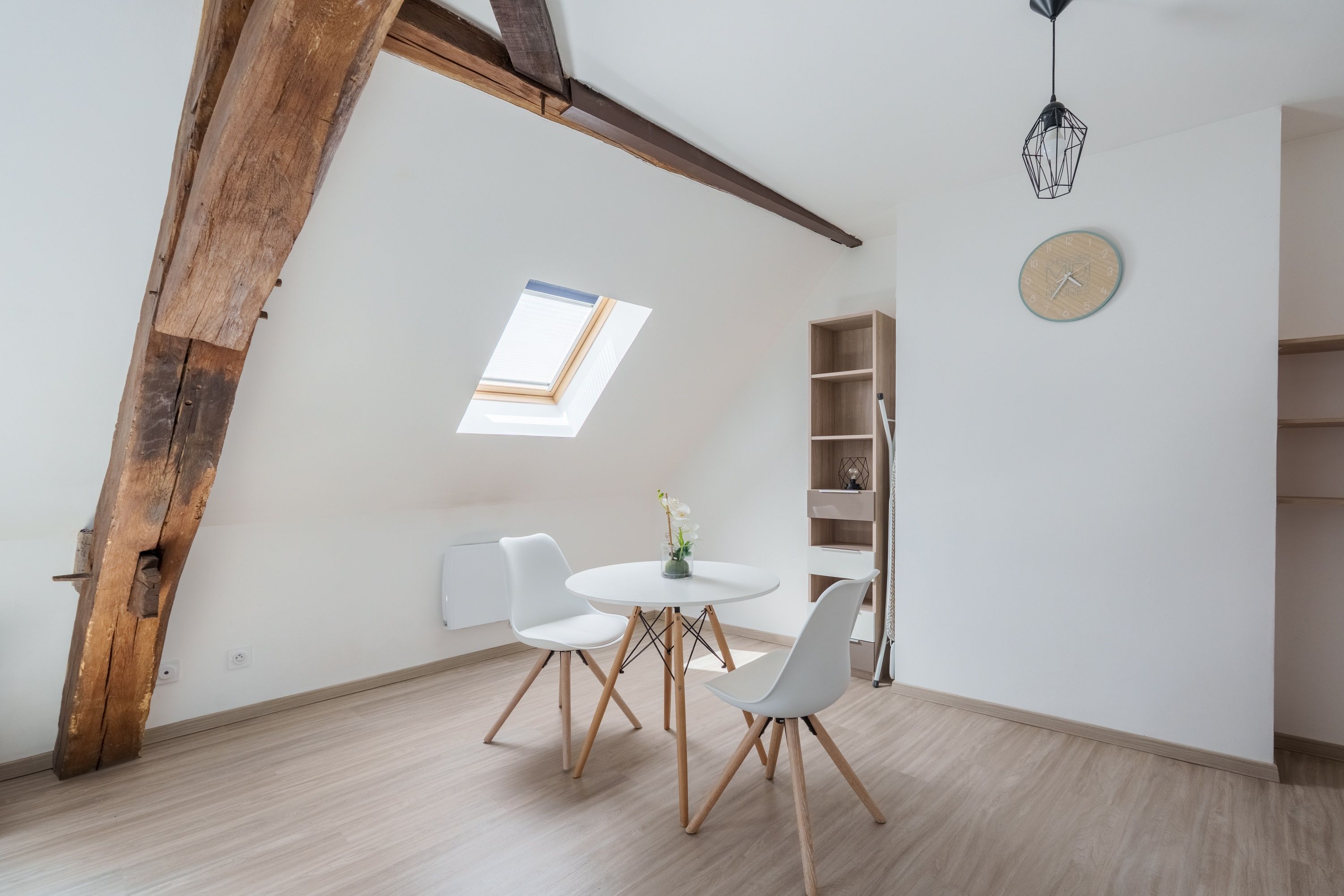 Une pièce avec un plafond en pente, un puits de lumière, une table ronde avec des chaises, une bibliothèque et une horloge murale.