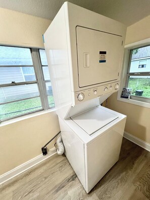 A compact, white, wall-mounted washer and dryer unit with a control panel featuring multiple dials and a small label. It is installed in a room with beige walls, a window providing a view of a residential area, and a wooden floor. - The Blue~nicorn Bungalow - 7 mins to DT (Jacksonville)