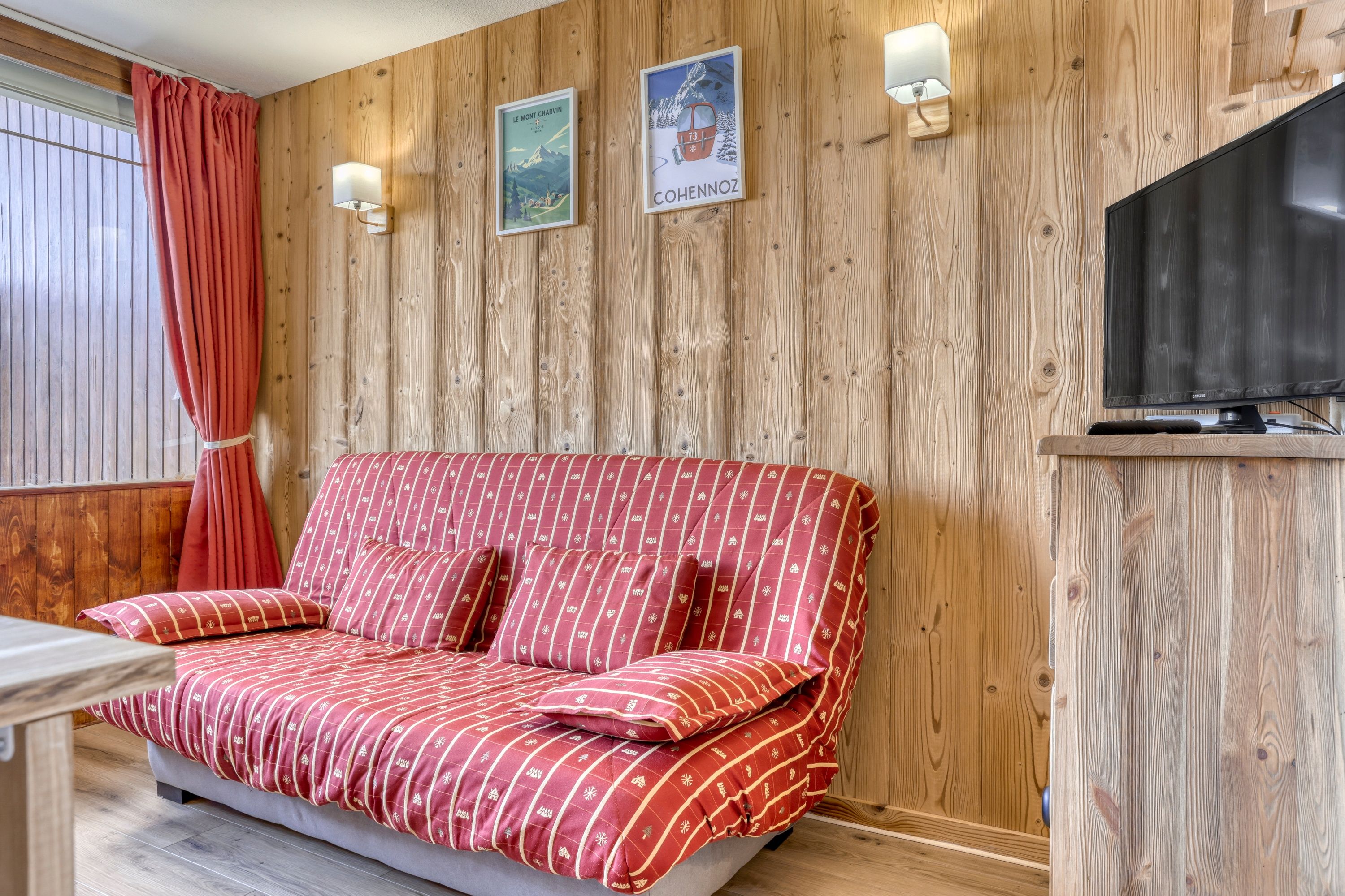 A cozy room with a wooden wall, a sofa with a red patterned cover, a television, and a window with curtains.