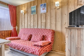 Ett mysigt rum med en väggpanel av trä, en soffa med ett rött, mönstrat överdrag, en tv och ett fönster med gardiner.