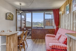 A room with a wooden floor, a red-striped sofa, a dining table with chairs, a window with a view of mountains, and a ceiling fan. - L’Écrin du Cernix - 400m du télésiège (Cohennoz)