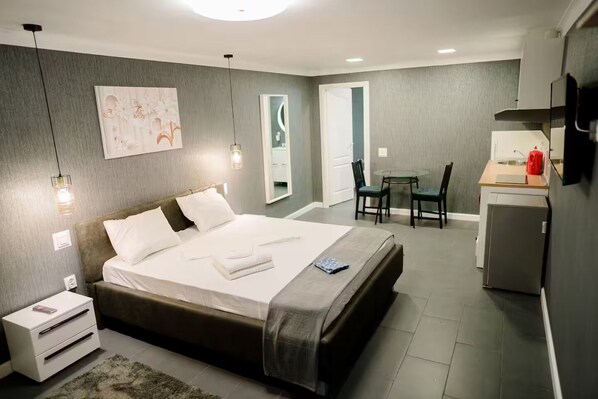 A modern hotel room with a large bed, bedside tables, a desk, and a refrigerator.