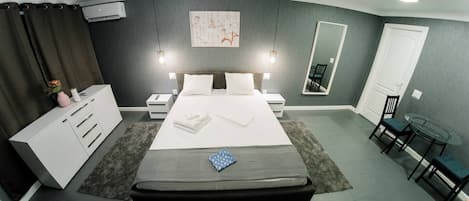 A modern hotel room with a large bed, bedside tables, a wardrobe, a small round table with chairs, and a mirror on the wall.