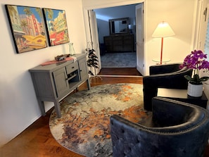 Ein Wohnzimmer mit einem dunkelbraunen Ledersessel, einer grauen Vitrine mit Weinflaschen, einem gemusterten Teppich und zwei Bildern an der Wand.