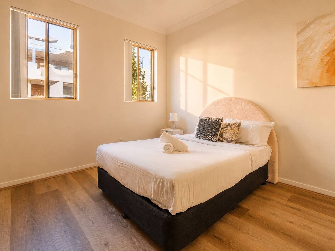 A bedroom with a bed, two windows, a nightstand, and a wall-mounted artwork.