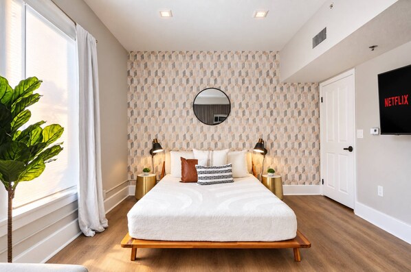 A modern bedroom with a large bed, two bedside tables, a round mirror, and a TV displaying 'NETFLIX'. - The Penn Hotel - Sleeps 3 & Gaslamp Nightlife Now! (San Diego)