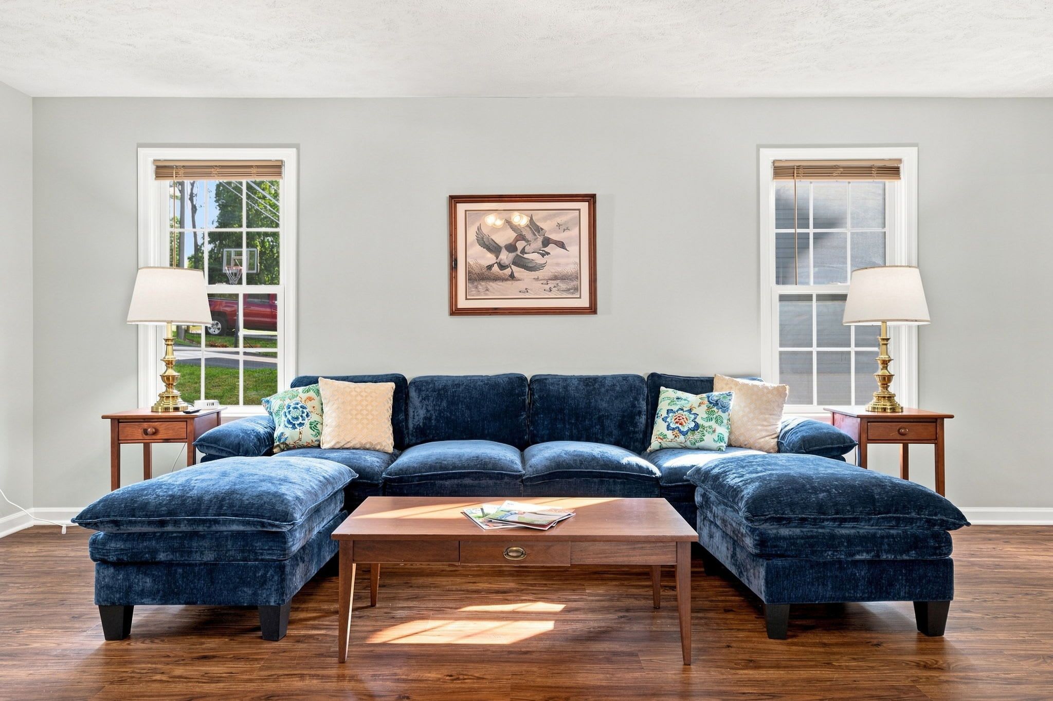 Uma sala de estar com um sofá seccional azul escuro, duas mesas laterais com abajures, um quadro emoldurado na parede e piso de madeira.