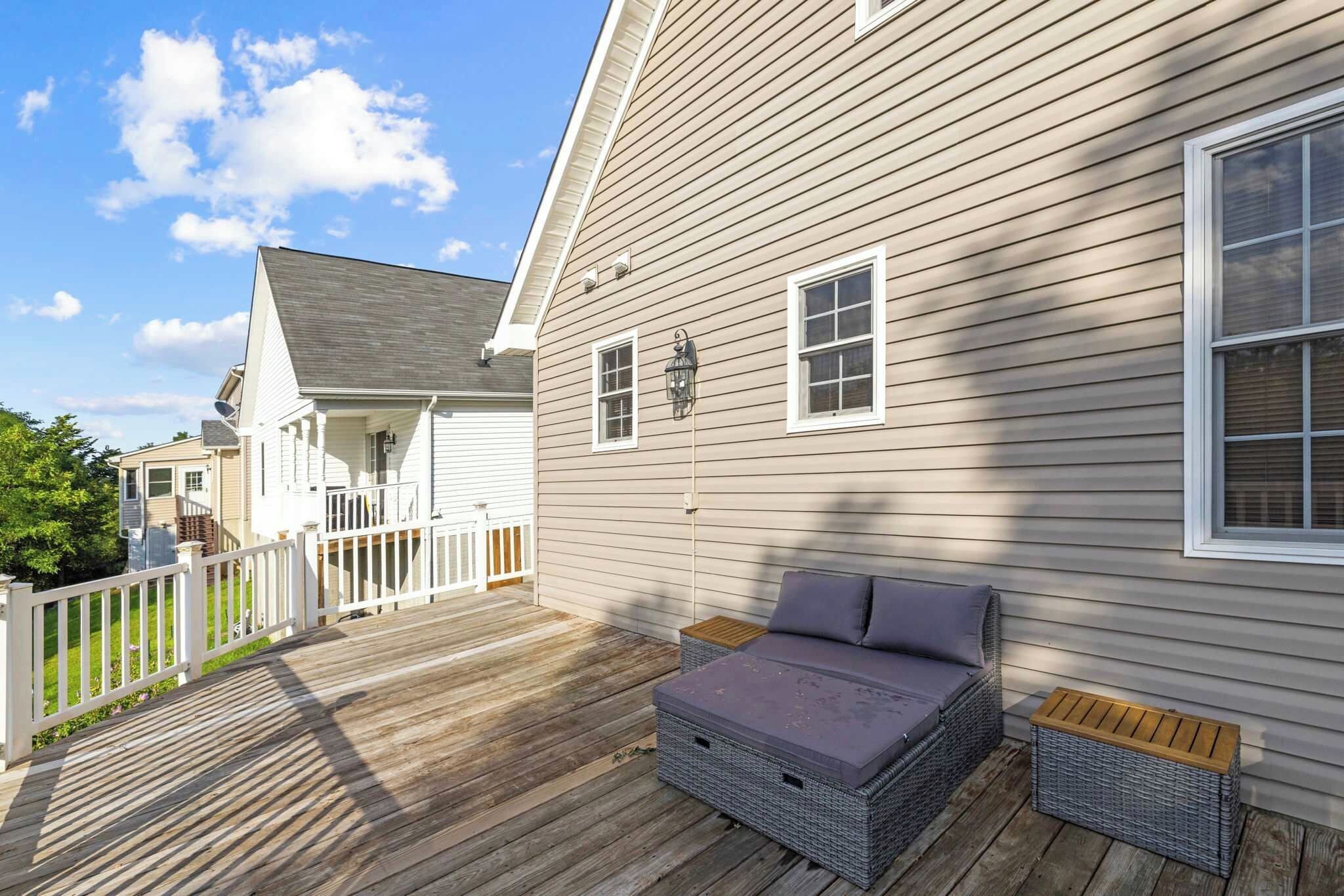 Une terrasse en bois avec un canapé en osier et une petite table, une clôture blanche et une maison aux murs recouverts d’un revêtement beige et dotée d’un toit gris.