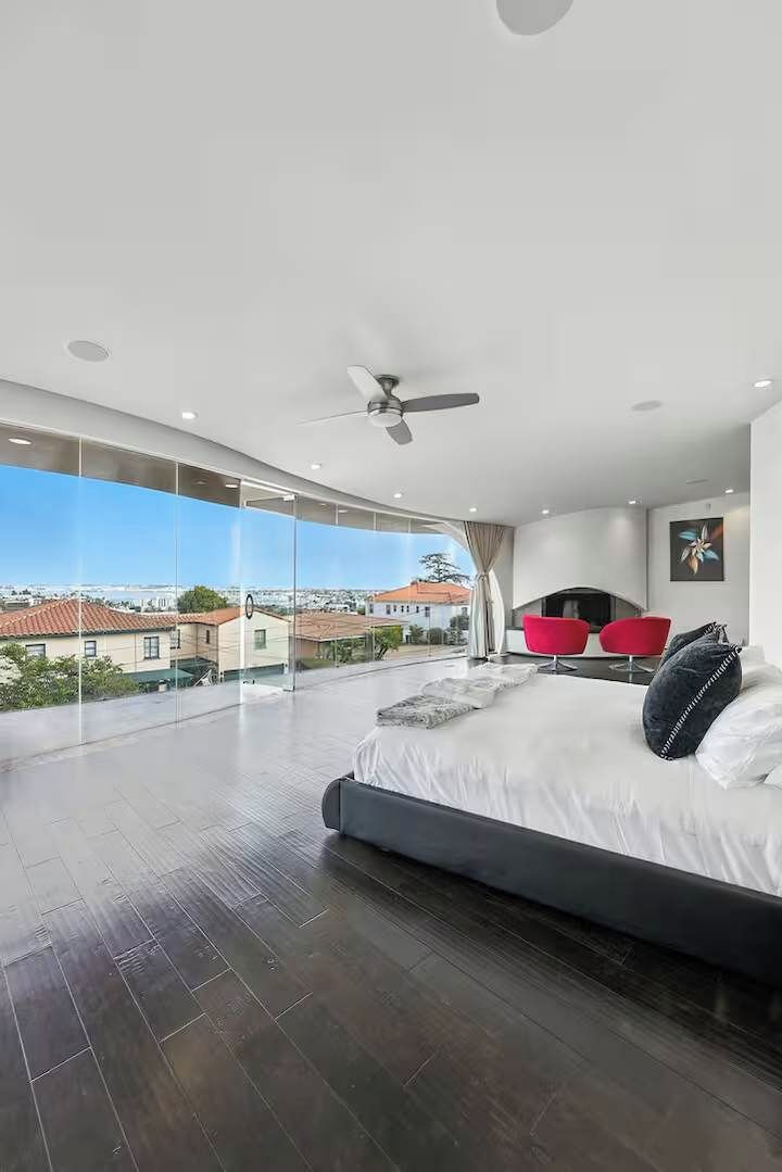 Une chambre moderne avec un grand lit, un ventilateur de plafond et une vue sur la ville grâce à de grandes fenêtres du sol au plafond.