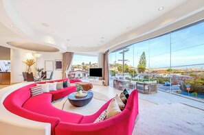 A modern living room with a large curved sofa, a coffee table, and a TV. There is a dining area with a table and chairs, and a large window offering a view of the city. - The Wave House!  Iconic Design - Panoramic Views (San Diego)